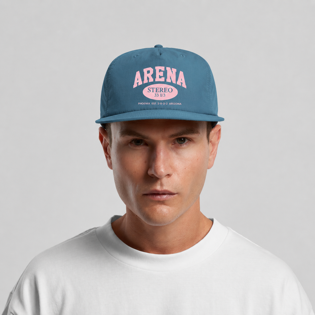 A man wearing a blue snapback cap with pink 'ARENA STEREO' text on the front, paired with a white t-shirt against a light gray background.