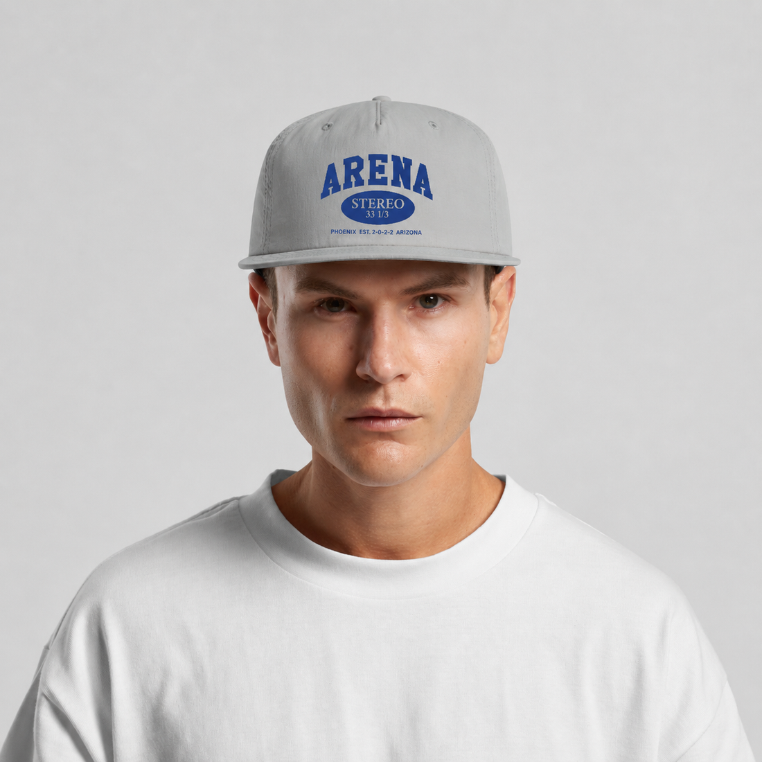 A man wearing a light gray snapback cap with blue 'ARENA' embroidery, paired with a white crewneck t-shirt, against a light gray background.