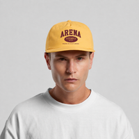 A man wearing a yellow snapback cap with a maroon 'ARENA STEREO' logo on the front, paired with a white t-shirt, against a light gray background.