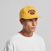 A man wearing a yellow snapback cap with a maroon 'ARENA STEREO' logo on the front, paired with a white t-shirt, photographed against a neutral light gray background.