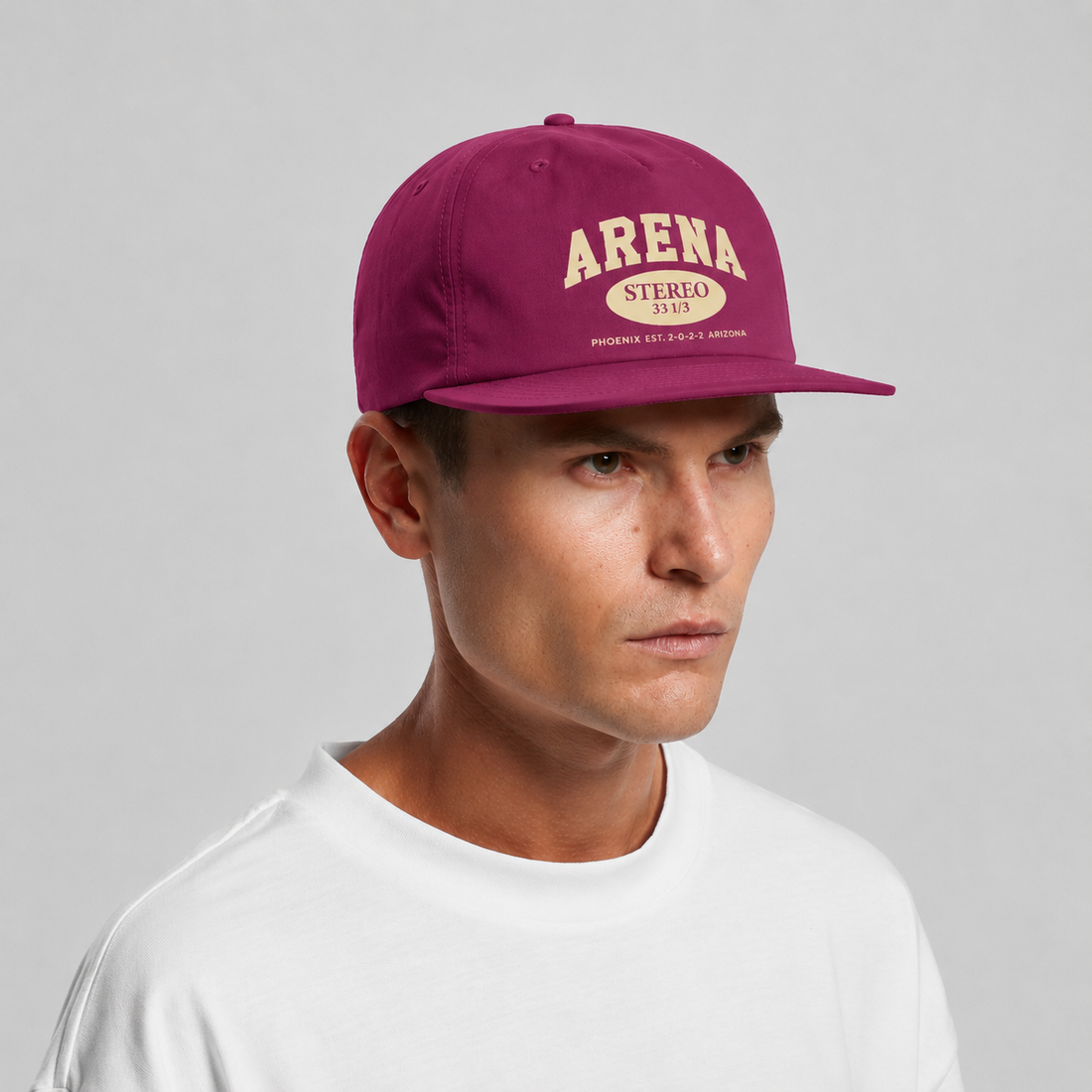 A man wearing a grape snapback cap with cream embroidered text reading 'ARENA STEREO' and a white t-shirt, photographed against a neutral gray background.