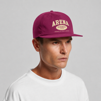 A man wearing a grape snapback cap with cream embroidered text reading 'ARENA STEREO' and a white t-shirt, photographed against a neutral gray background.