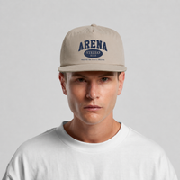 A man wearing a beige snapback cap with navy blue 'ARENA STEREO' text on the front, paired with a white t-shirt against a light gray background.