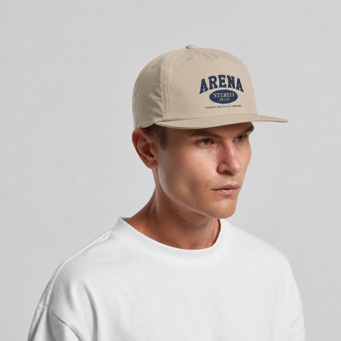 A man wearing a beige snapback cap with a navy blue 'ARENA STEREO' logo on the front, paired with a white t-shirt, against a light gray studio background.