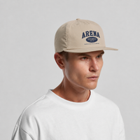A man wearing a beige snapback cap with a navy blue 'ARENA STEREO' logo on the front, paired with a white t-shirt, against a light gray studio background.