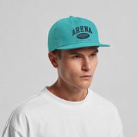 A man wearing a turquoise snapback cap with a dark navy 'ARENA STEREO' graphic on the front, paired with a white t-shirt, against a light gray studio background.