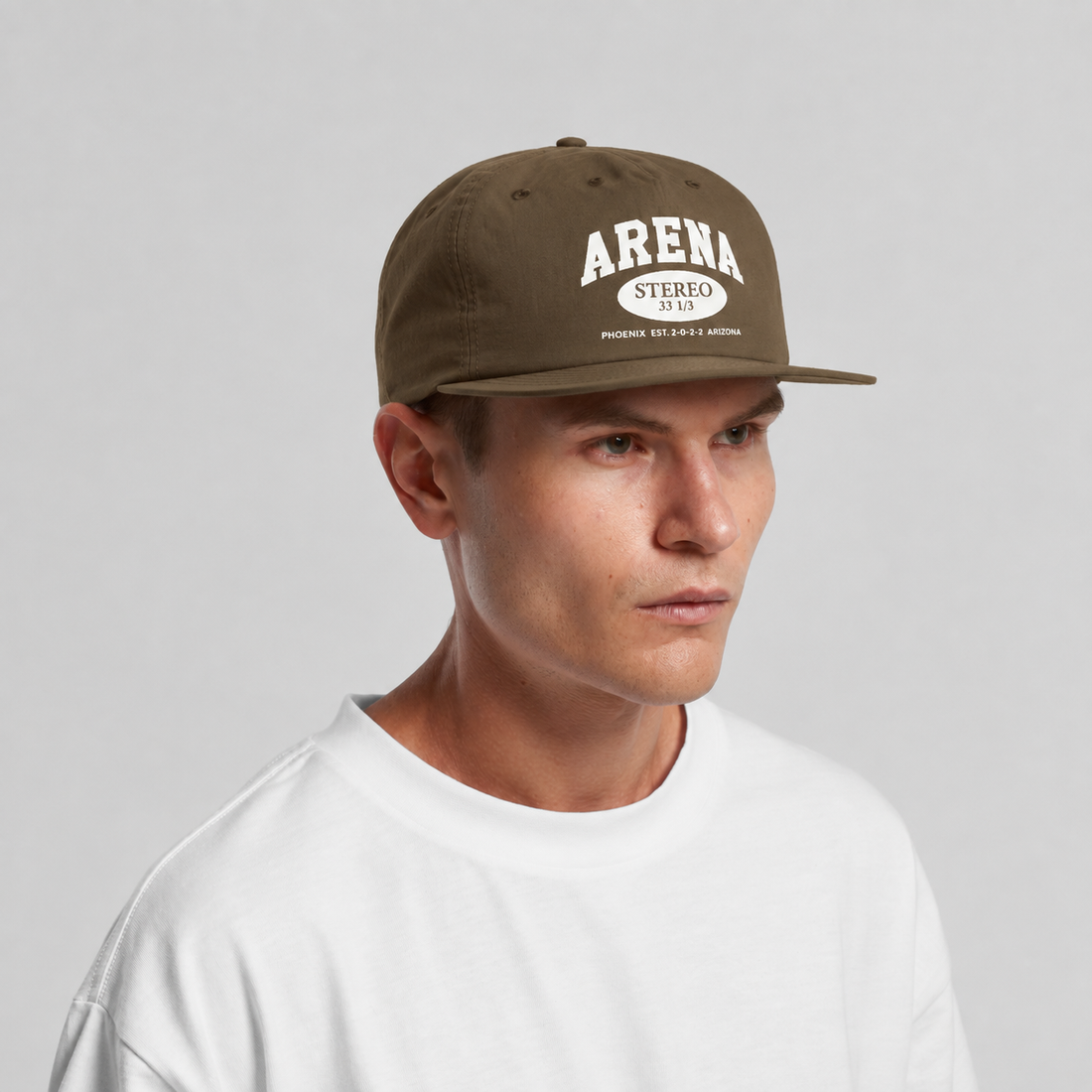 A man wearing a brown snapback cap with 'ARENA STEREO' printed in white on the front, paired with a white t-shirt, against a light gray background.