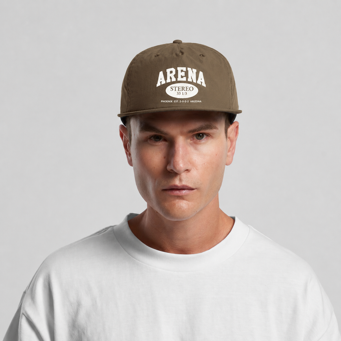 A man wearing a brown snapback cap with 'ARENA STEREO' printed in white on the front, paired with a white t-shirt, against a light gray background.