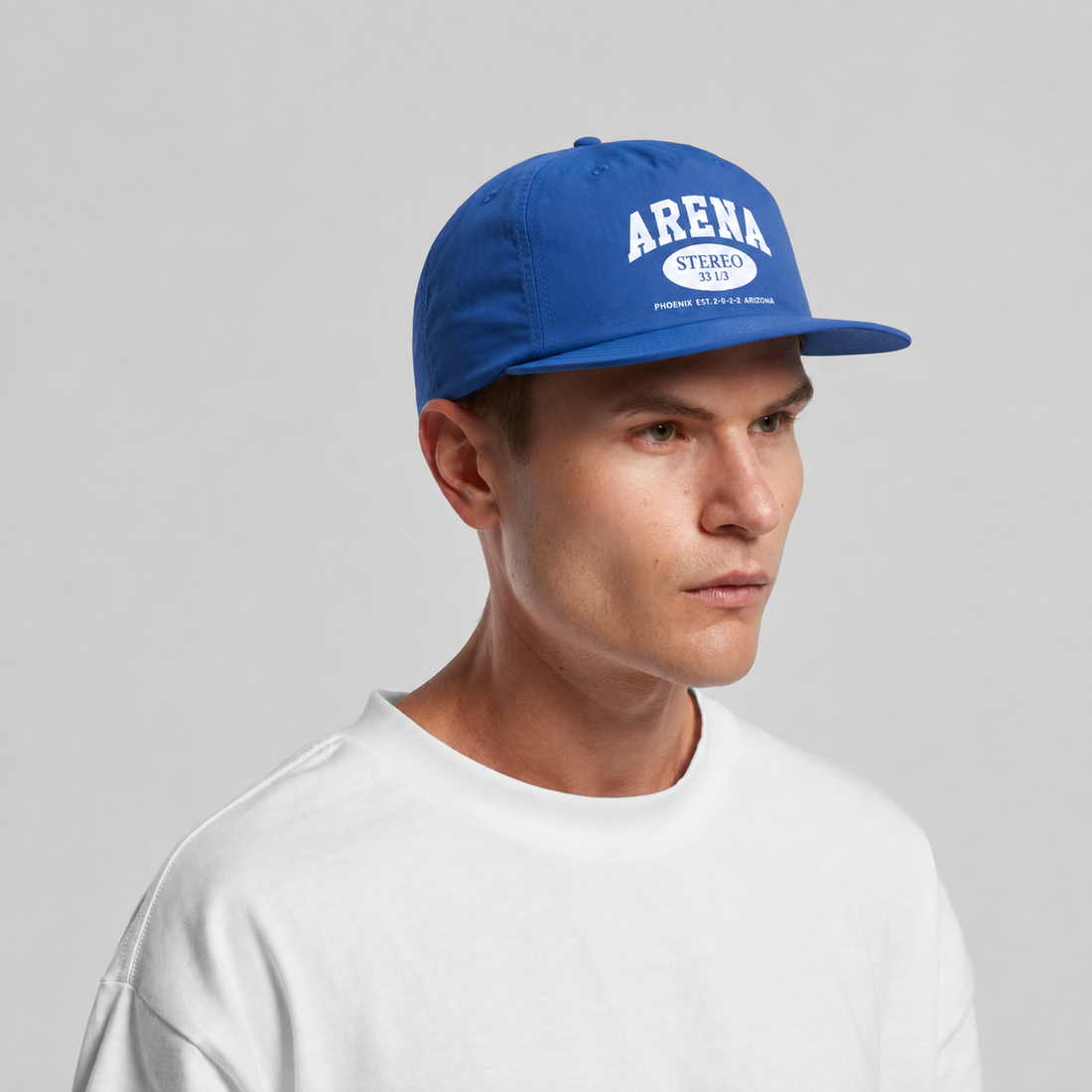 A man wearing a royal blue snapback cap with 'ARENA STEREO' printed in white on the front, paired with a white t-shirt, against a light gray studio background.