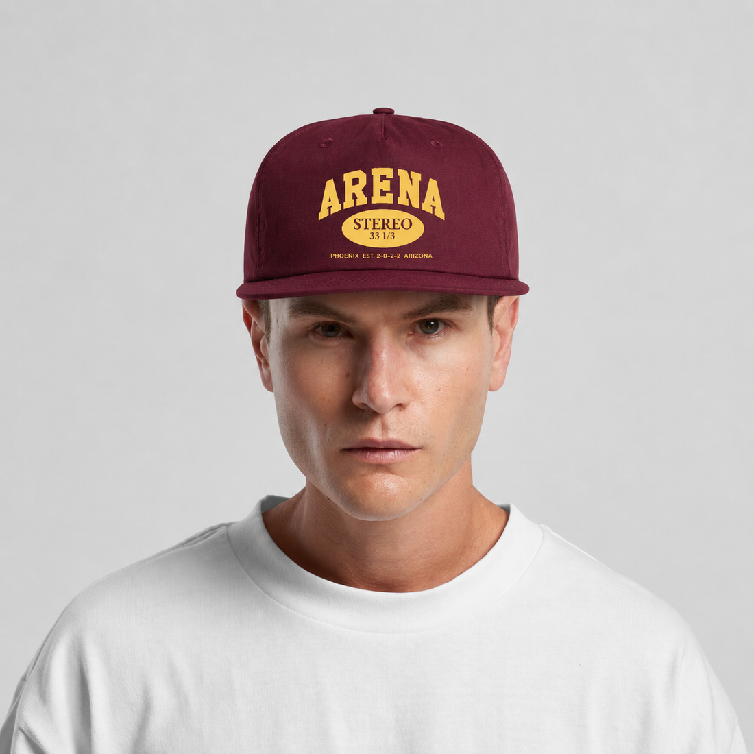 A man wearing a maroon snapback cap with yellow embroidered text reading 'ARENA STEREO' and a white crewneck t-shirt, photographed against a light gray studio background.
