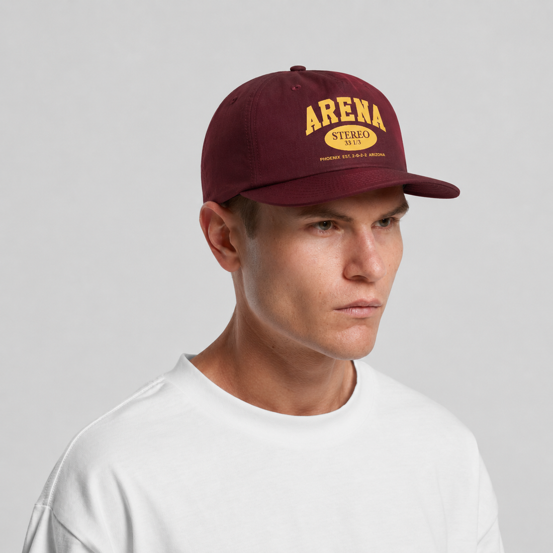 A man wearing a maroon baseball cap with yellow 'ARENA STERO' on the front, paired with a white t-shirt, against a light gray background.