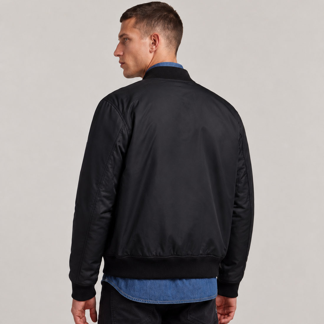 A man shown from the back wearing a black bomber jacket over a blue shirt and black pants, photographed against a plain light gray studio background.