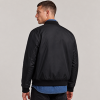 A man shown from the back wearing a black bomber jacket over a blue shirt and black pants, photographed against a plain light gray studio background.