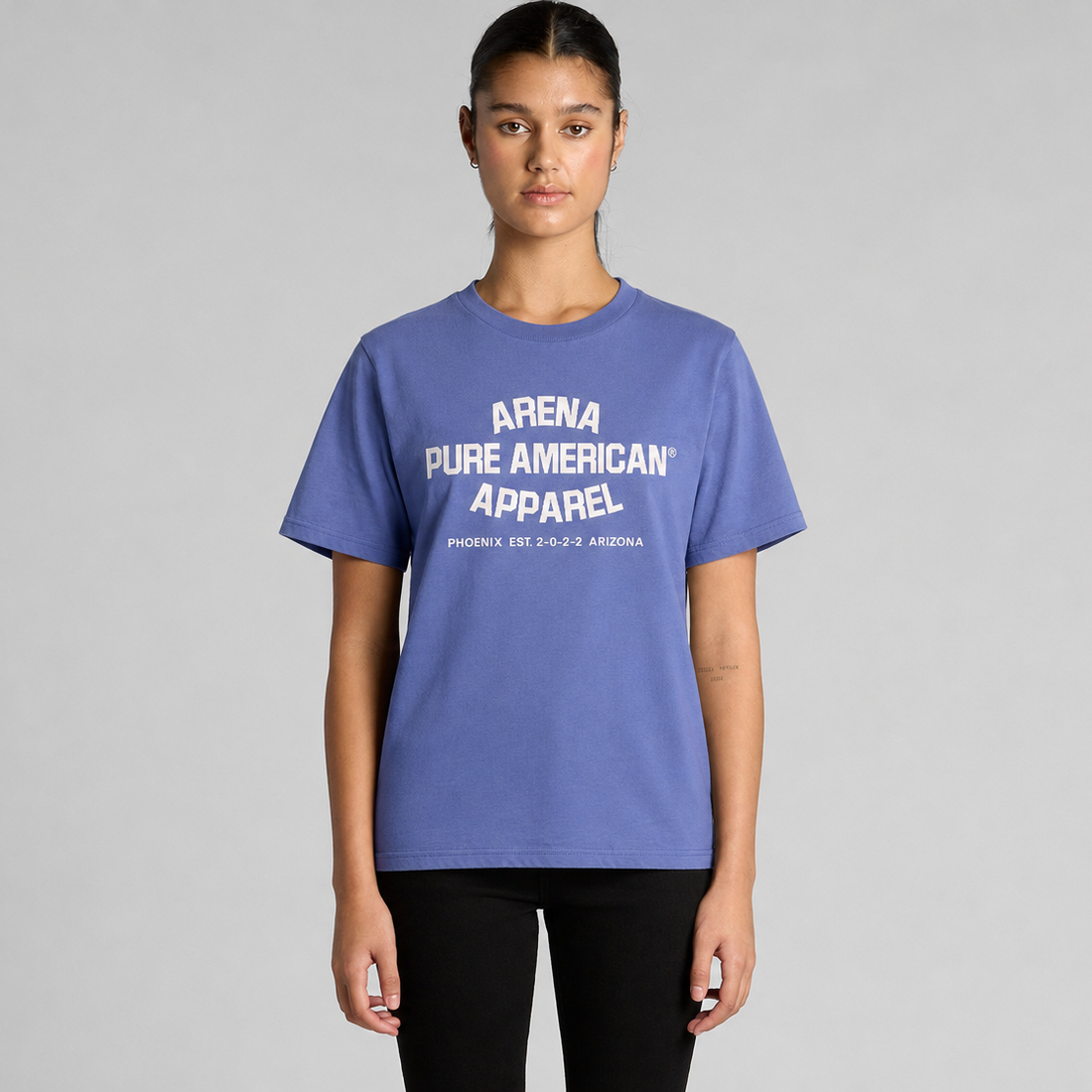 A model wearing a lapis blue crewneck t-shirt with white text reading 'ARENA PURE AMERICAN APPAREL' on the chest, paired with black pants, against a solid gray studio background.