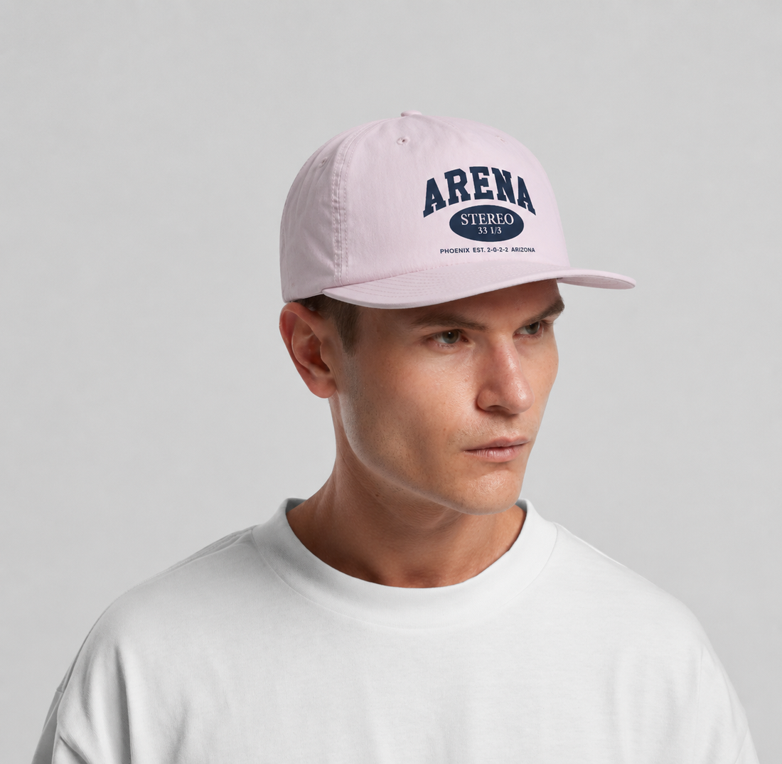 A man wearing a orchid pink baseball cap with navy blue 'ARENA STEREO' text on the front, paired with a white crewneck t-shirt, against a solid light gray background.
