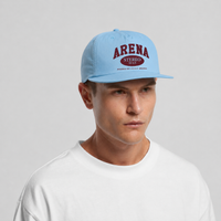 A man wearing a light blue snapback cap with a maroon 'ARENA STEREO' logo on the front, paired with a white t-shirt against a light gray background.