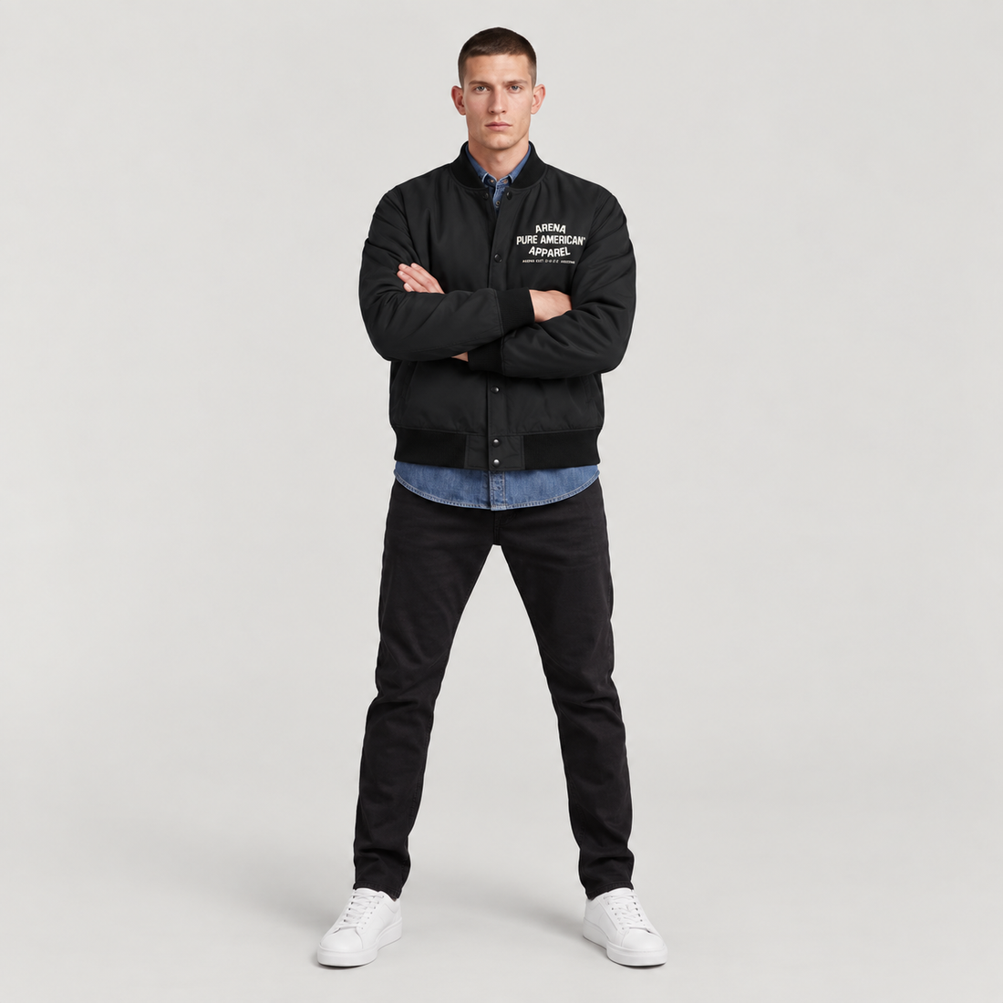 A man modeling a black varsity-style jacket with white embroidered text on the left chest, worn over a blue button-up shirt and paired with black pants and white sneakers, against a light gray studio background.