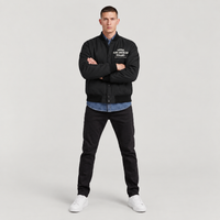 A man modeling a black varsity-style jacket with white embroidered text on the left chest, worn over a blue button-up shirt and paired with black pants and white sneakers, against a light gray studio background.