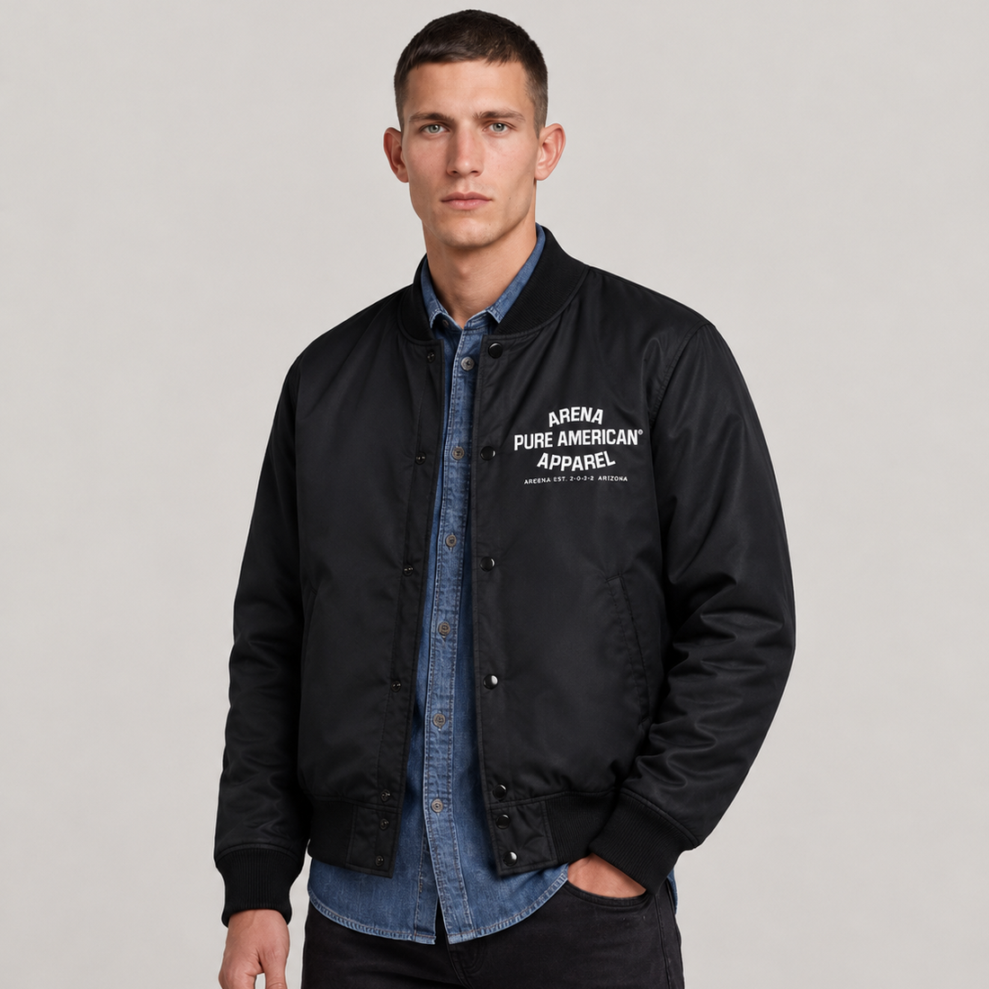 A man wearing a black Arena Pure American Apparel bomber jacket over a blue denim shirt and black pants, standing against a light gray studio background.