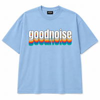 A light blue short-sleeve t-shirt with a retro-style graphic on the chest reading 'goodnoise' in white letters with a white outline, layered over vertical rainbow stripes in red, orange, yellow, green, and blue. The brand name 'ARENA' is visible on the inner neck label.