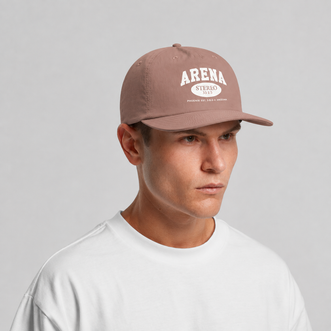 A man wearing a dusty rose baseball cap with 'ARENA STEREO' printed in white on the front, paired with a white t-shirt against a light gray background.
