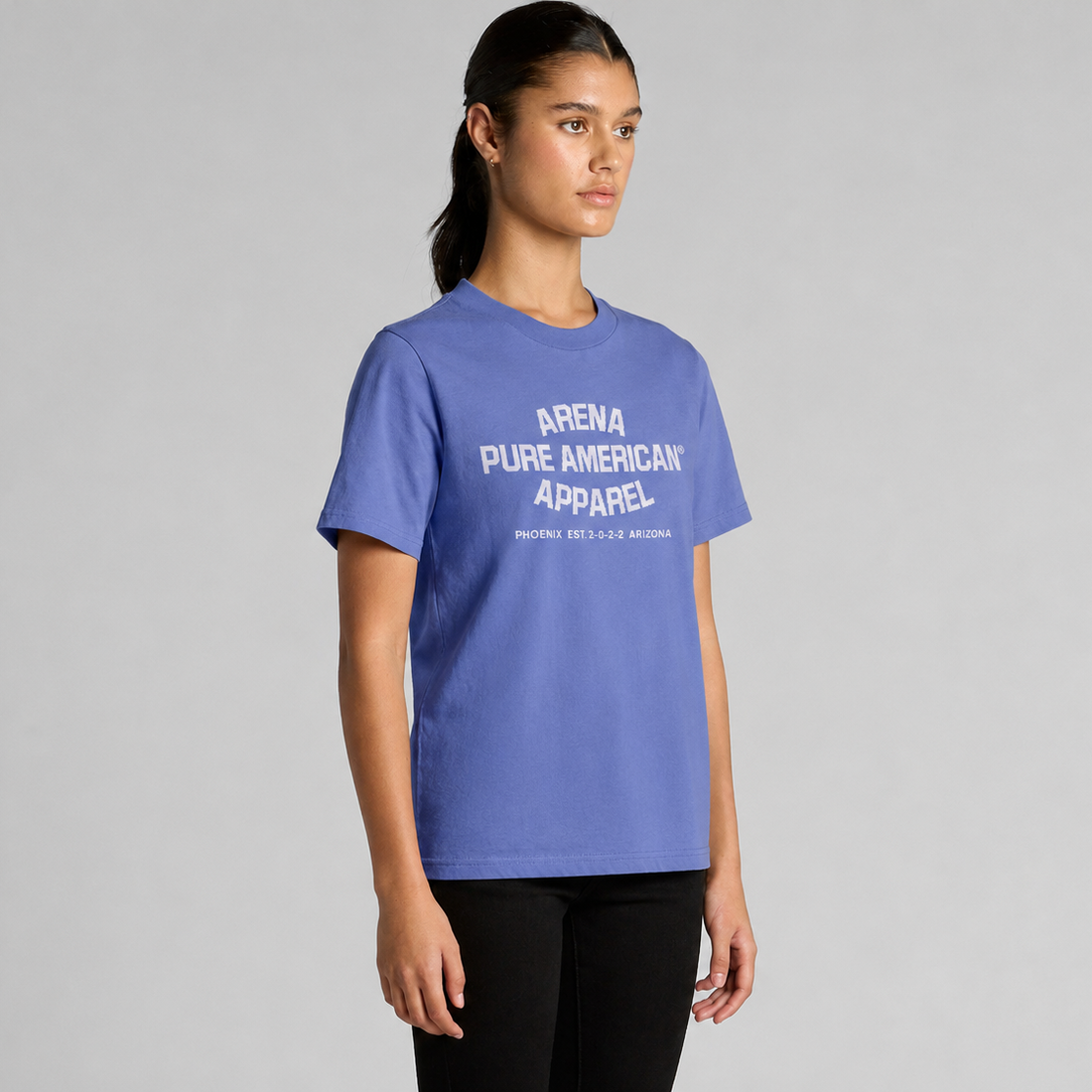 A woman modeling a lapis blue short-sleeve t-shirt with white text reading 'ARENA PURE AMERICAN APPAREL' on the chest, set against a plain gray studio background.