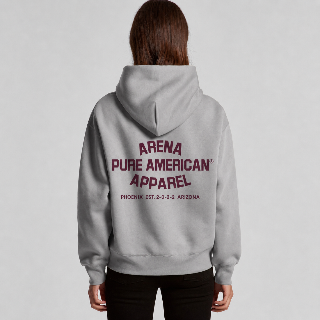 A woman viewed from the back wearing a light gray pullover hoodie with 'ARENA PURE AMERICAN APPAREL' printed in maroon across the back, paired with black pants, against a light gray studio background.
